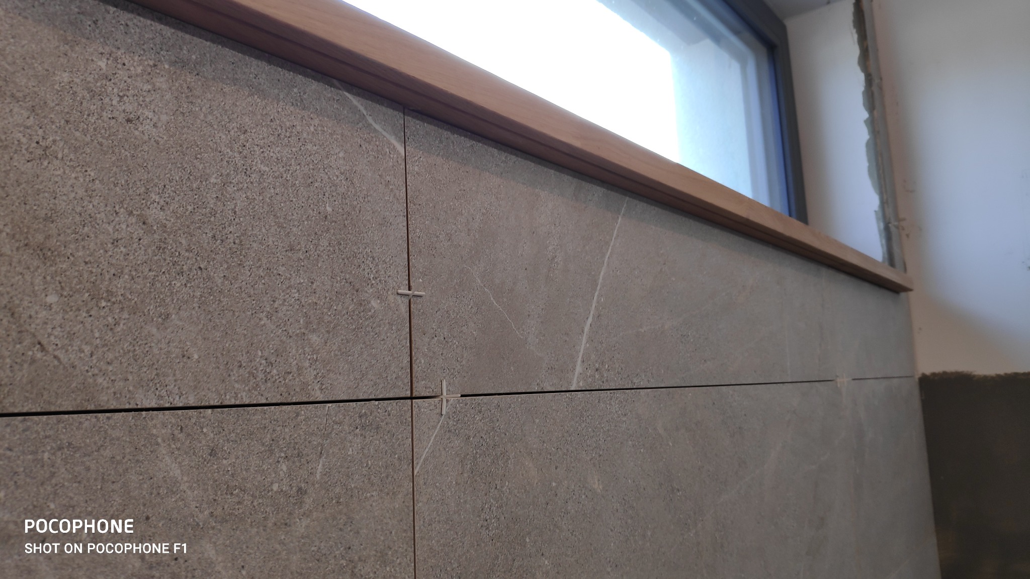 Układanie szarych płytek ceramicznych przy oknie z drewnianym parapetem, krzyżyki dystansowe w fugach, widoczne ślady zaprawy na ścianie.