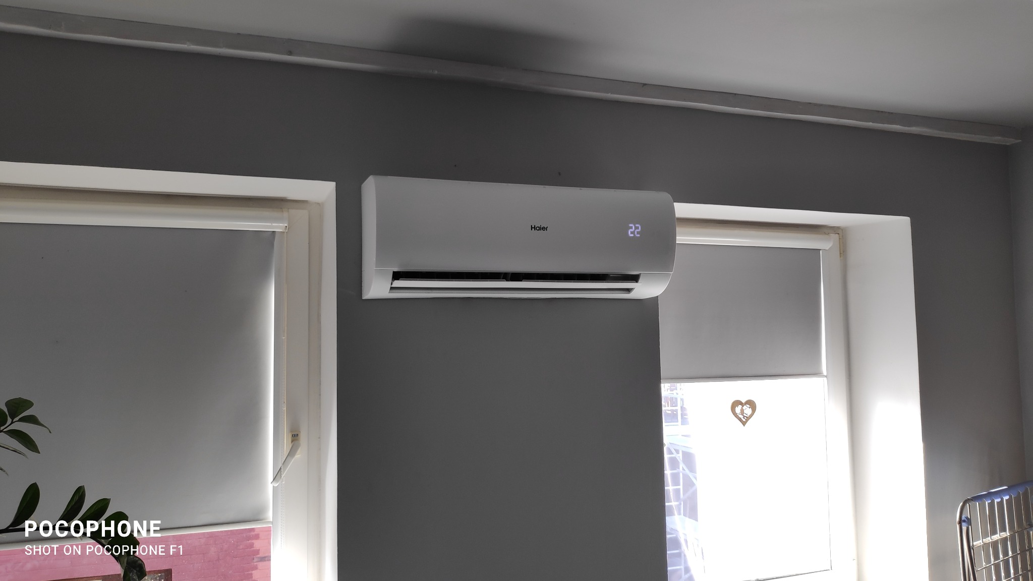 Biały klimatyzator Haier zamontowany na szarej ścianie w pokoju, widoczne rolety w oknach po bokach.
