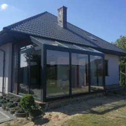 Szklana oranżeria z czarnymi ramami, przylegająca do domu z ciemną dachówką, widok z zewnątrz w słoneczny dzień.