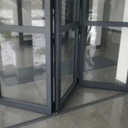 Rozkładane drzwi balkonowe z szarymi ramami, widoczne prowadnice podłogowe i fragment wnętrza z rośliną.