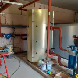 Instalacja centralnego ogrzewania z dużym zbiornikiem buforowym, rurami w kolorze ceglastym i osprzętem, widoczne puszki z farbą i narzędzia na pierwszym planie.