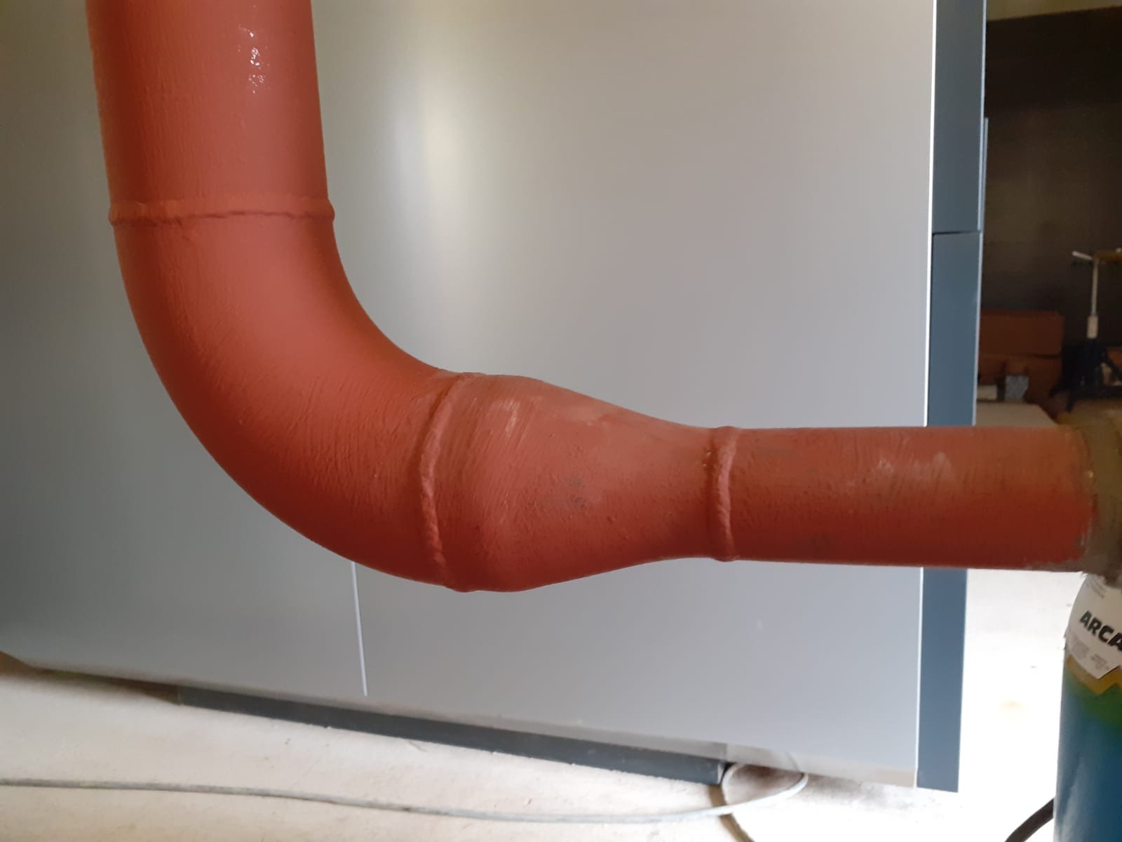 Czerwona, spawana rura z kolankiem, pomalowana farbą, widoczna na tle szarej ściany i podłogi.