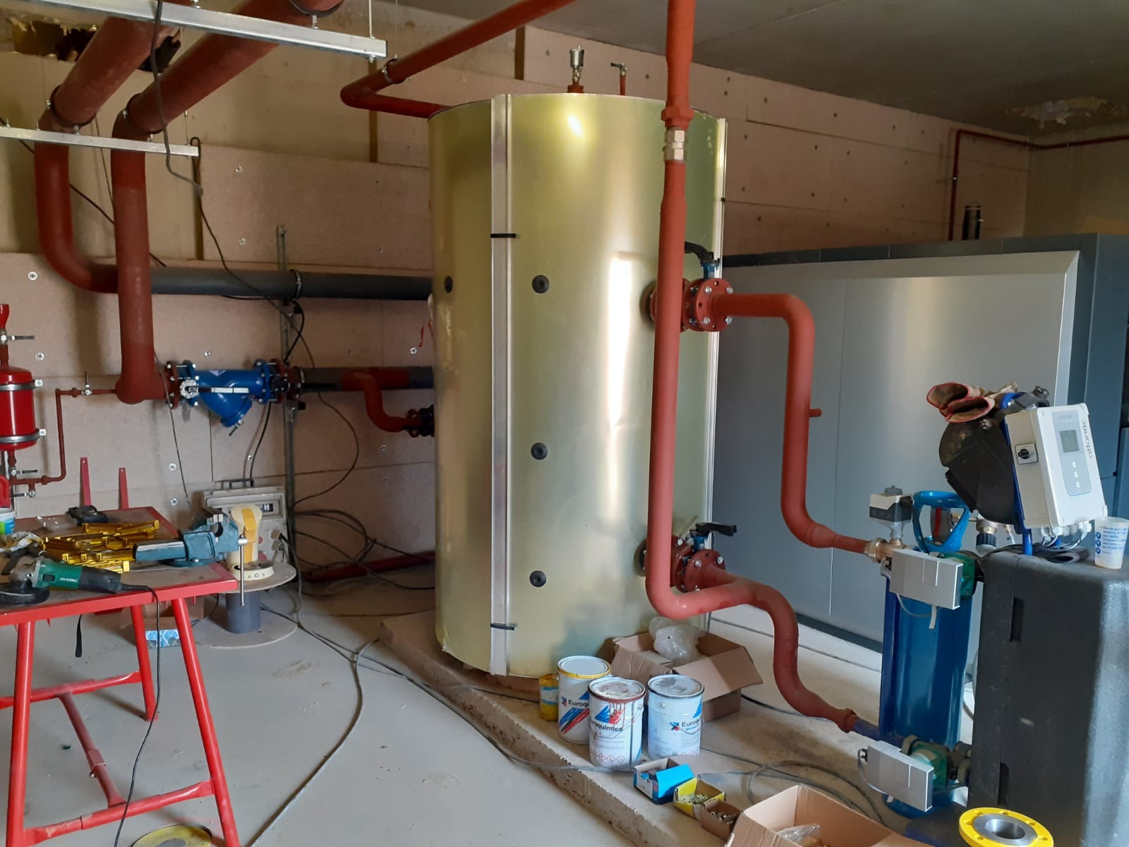 Instalacja centralnego ogrzewania z dużym zbiornikiem buforowym, rurami w kolorze ceglastym i osprzętem, widoczne puszki z farbą i narzędzia na pierwszym planie.