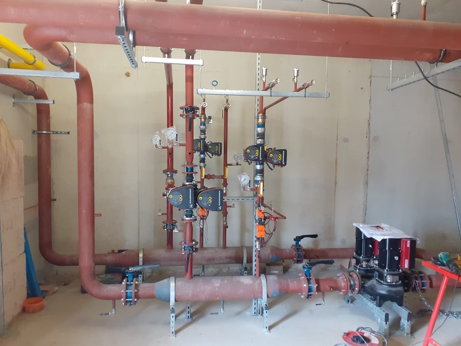 Instalacja centralnego ogrzewania z rurami w kolorze ceglastym, zaworami, pompami obiegowymi Grundfos i manometrami, zamontowana w surowym pomieszczeniu.