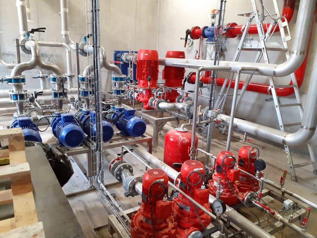 Kompleksowy system rurociągów z pompami w kolorach czerwonym i niebieskim, zainstalowany w surowym wnętrzu budynku, widoczne drabiny i elementy konstrukcyjne.