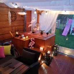 Zadaszony taras z drewnianą podłogą i meblami ogrodowymi, oświetlony lampionami i lampkami, widok na ogród z huśtawką.