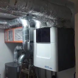 Wnętrze kotłowni z rekuperatorem Aeris Next, widoczne przewody wentylacyjne w izolacji, filtr powietrza w pomarańczowej ramie i drabina oparta o ścianę.