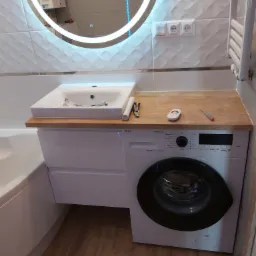 Nowoczesna łazienka z umywalką nablatową na szafce z pralką, okrągłe lustro z podświetleniem LED, płytki ścienne z fakturą, drewniany blat i narzędzia na nim.