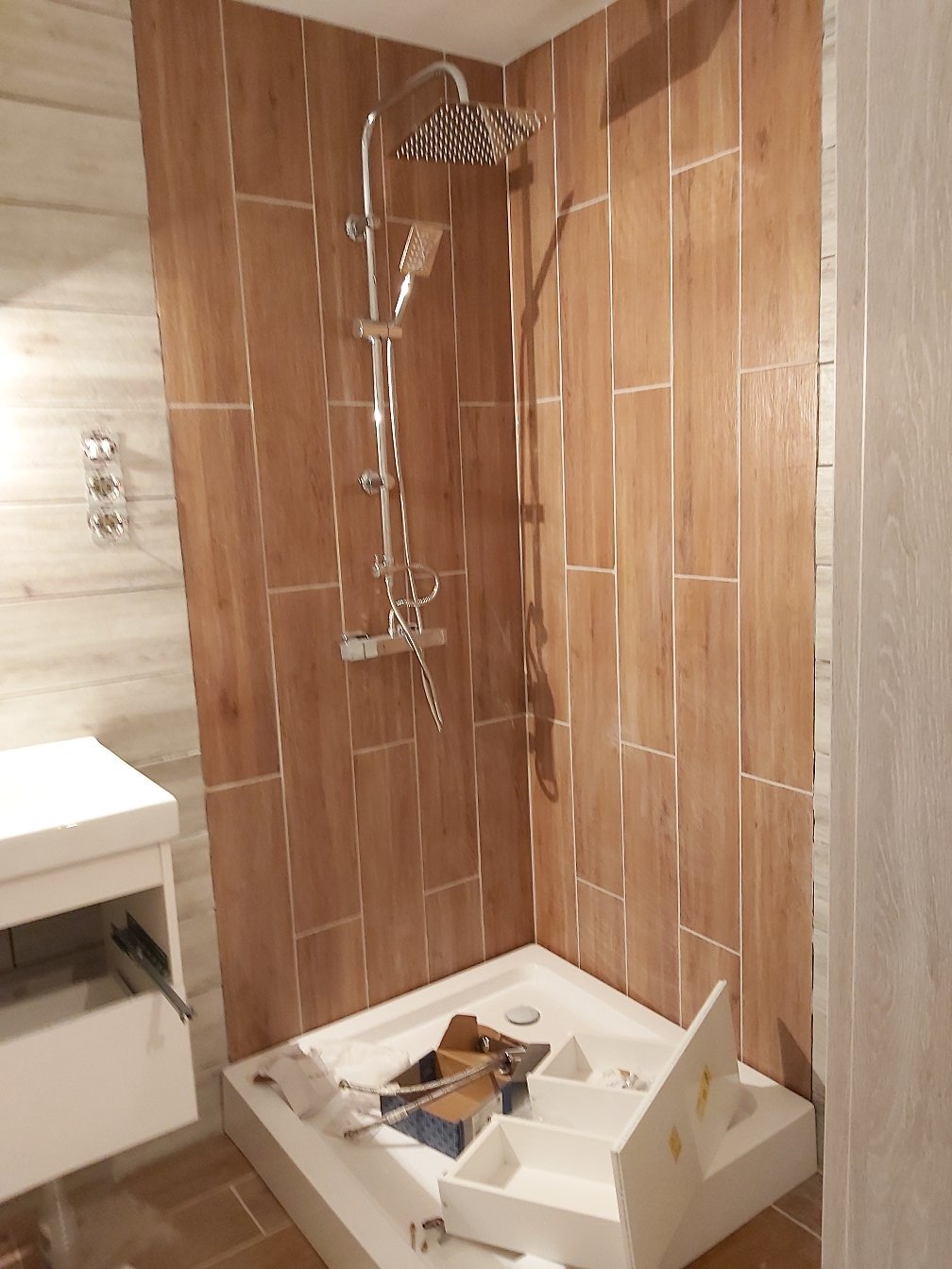 Montaż kabiny prysznicowej z deszczownicą i baterią termostatyczną na tle płytek imitujących drewno, widoczne elementy instalacji i biała szafka pod umywalkę z wysuniętą szufladą.