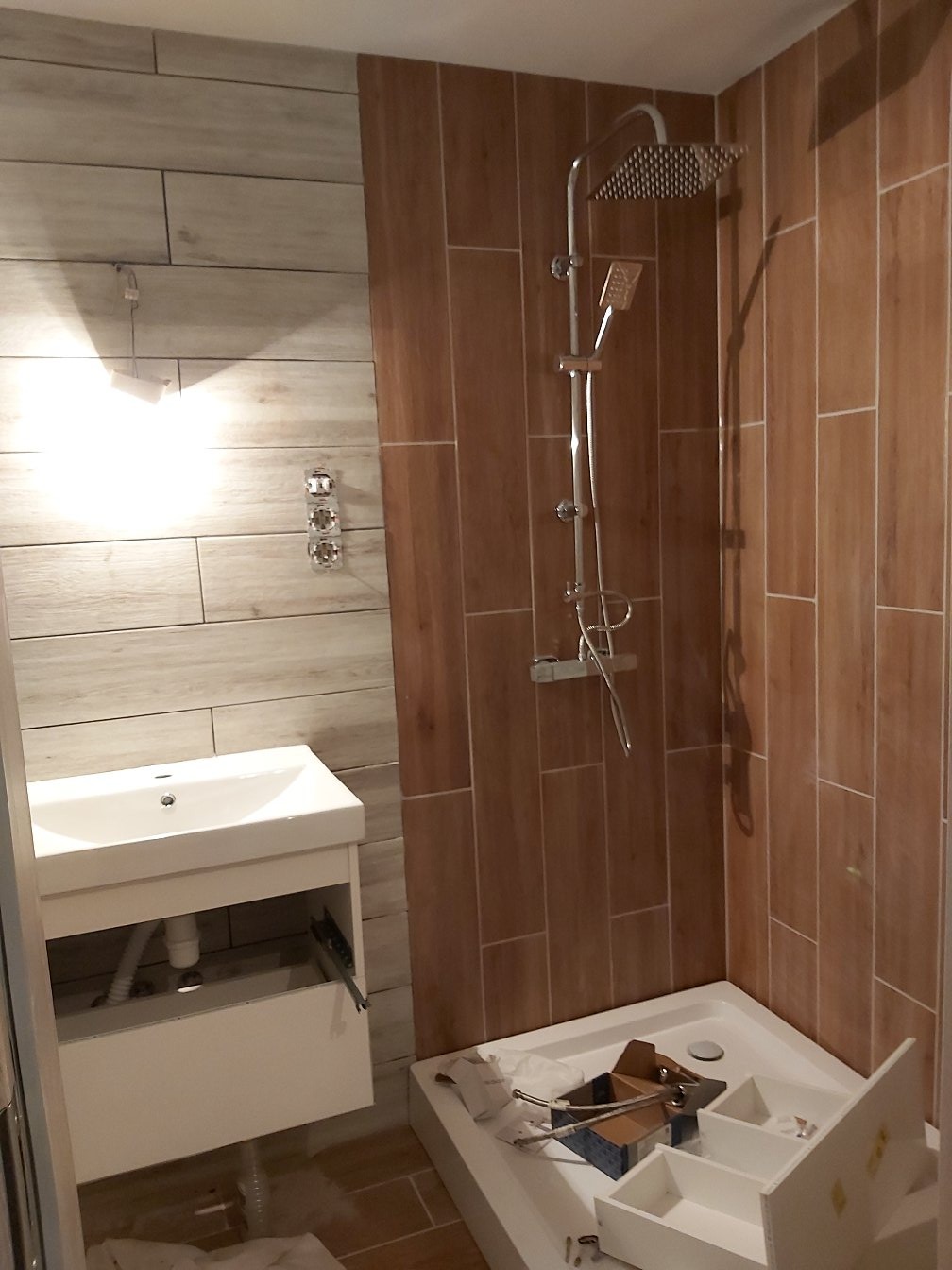 Niedokończona łazienka z drewnopodobnymi płytkami na ścianach, biała umywalka z otwartą szufladą, brodzik z otwartymi pudełkami i elementami armatury, chromowana bateria prysznicowa z deszczownicą.