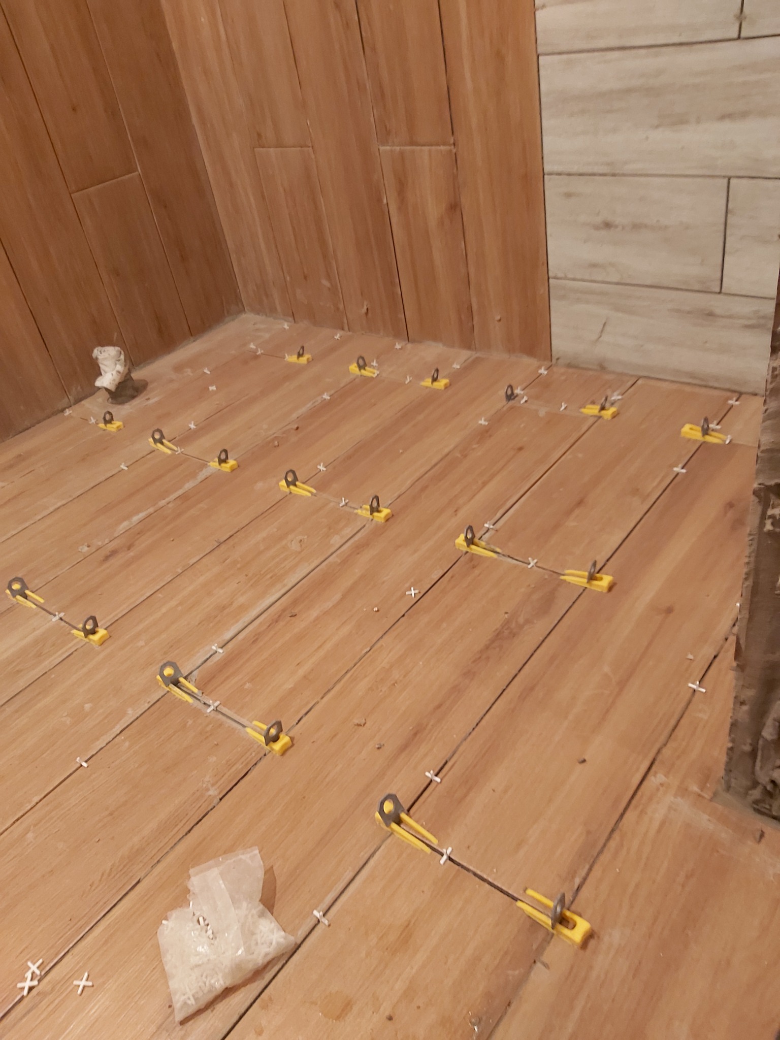 Układanie płytek imitujących drewno na podłodze, widoczne żółte klipsy poziomujące i krzyżyki dystansowe, w tle fragment ściany z jasnymi płytkami.