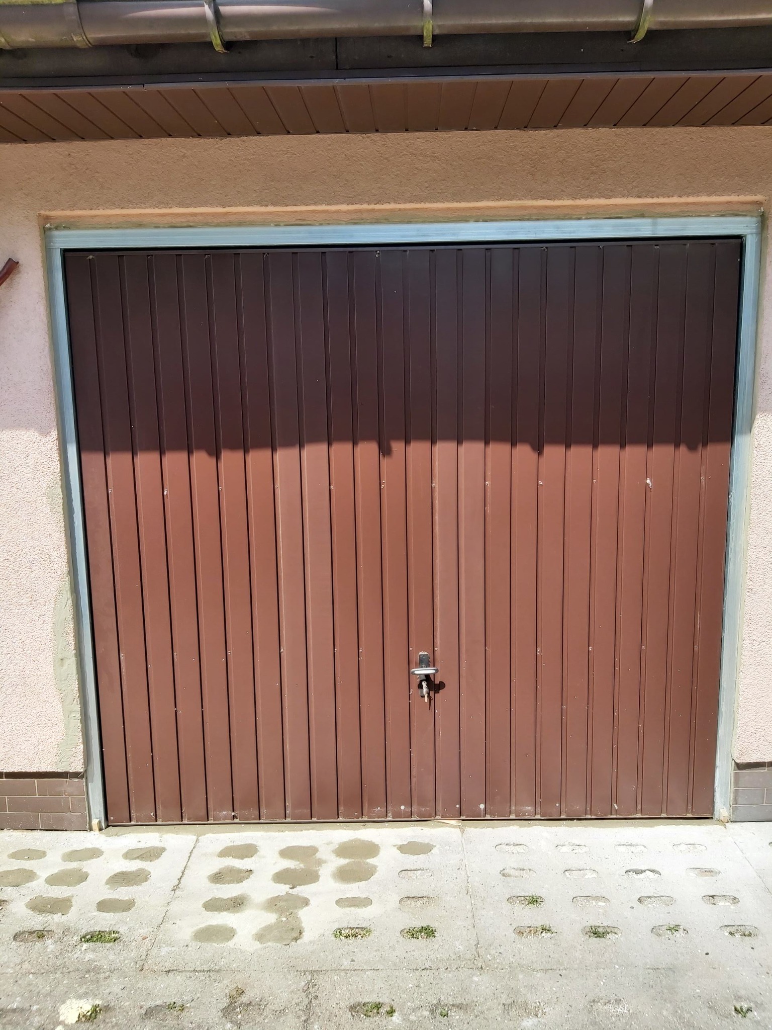 Brązowa brama garażowa segmentowa z widocznym zamkiem, osadzona w jasnoróżowej ścianie z szarą ramą, na tle betonowej nawierzchni z ażurowymi płytami.