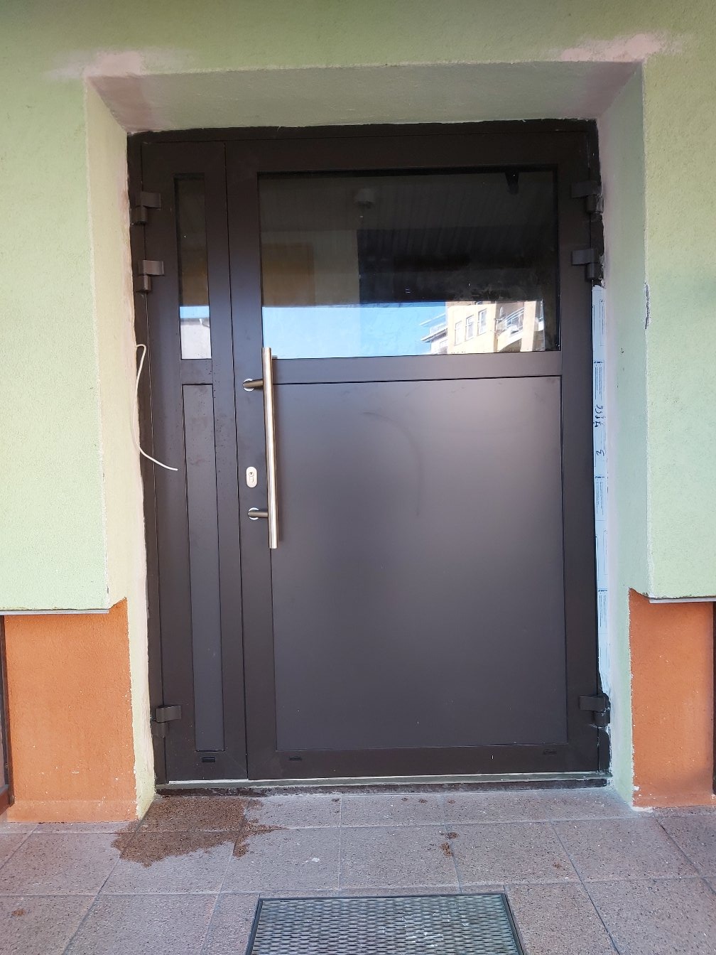 Ciemna, metalowa brama wejściowa z bocznym przeszkleniem, widoczne ślady montażu przy podłożu, obok fragment elewacji w dwóch kolorach.