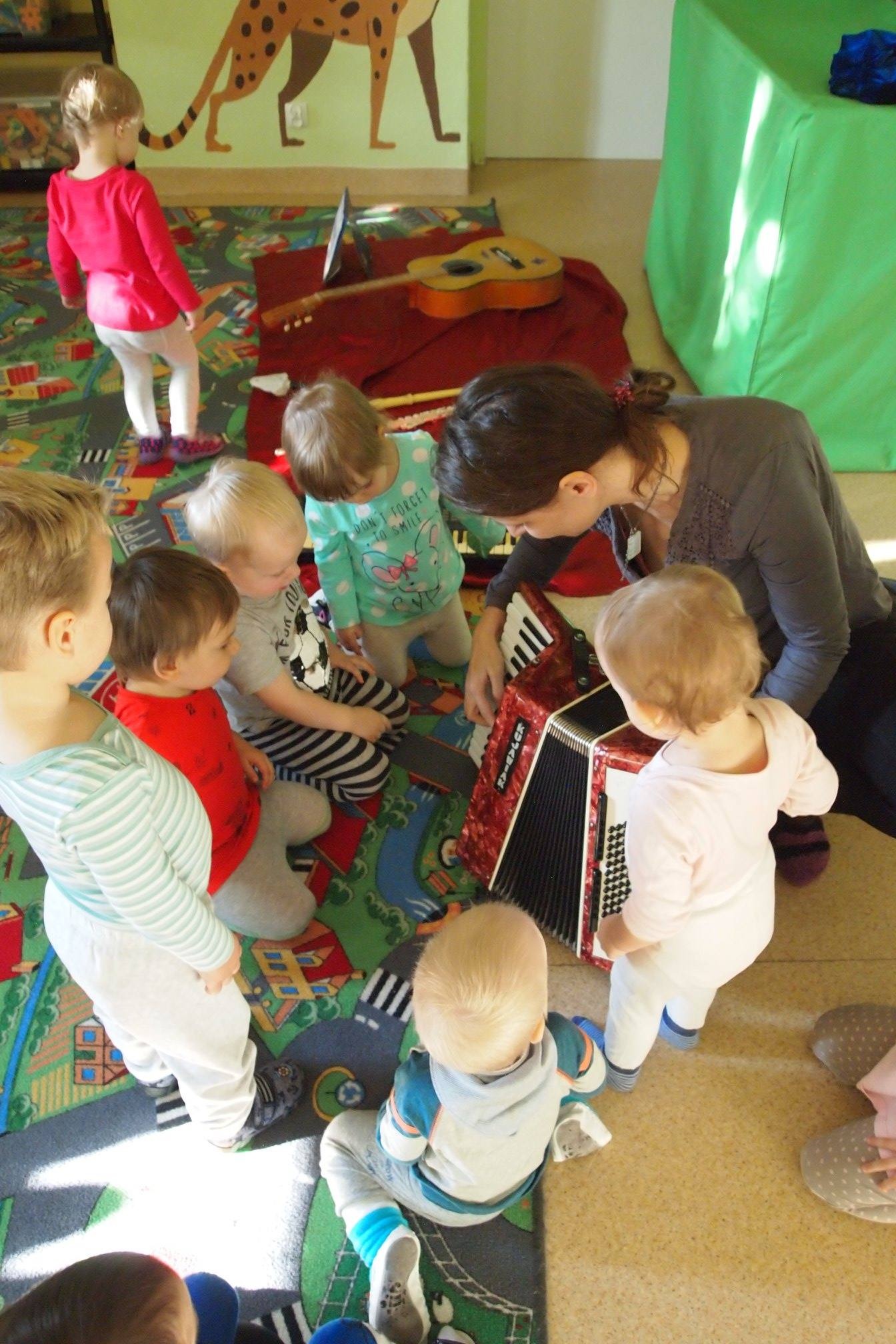 Animator gra na akordeonie otoczony grupą małych dzieci siedzących na kolorowym dywanie z motywem ulic i domów, w tle gitara i dekoracja ścienna z gepardem.