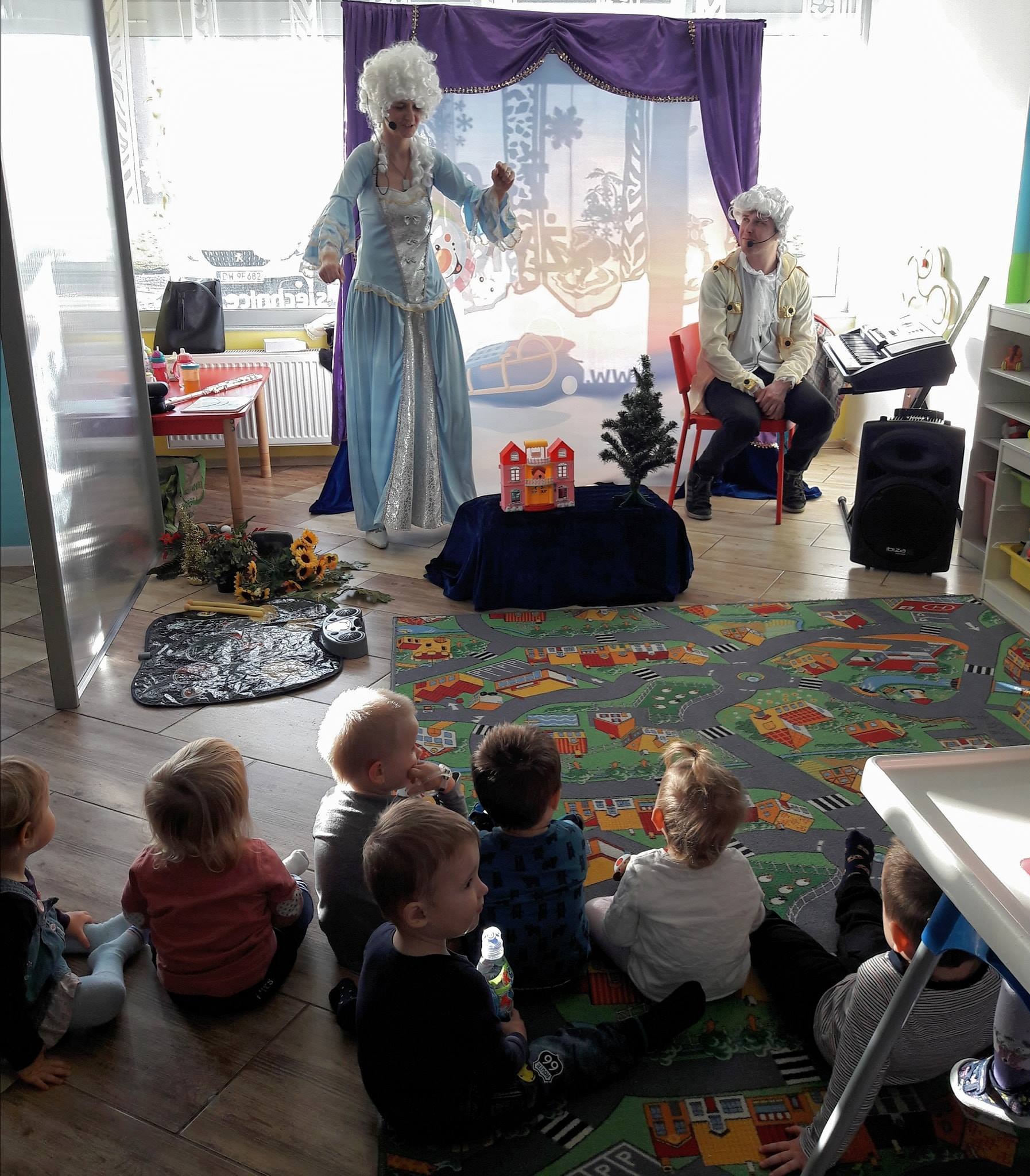 Dwoje aktorów w kostiumach z epoki, perukach i mikrofonach nagłownych występuje przed grupą dzieci siedzących na dywanie z motywem miasteczka. W tle scenografia z motywami zimowymi i fioletowe...