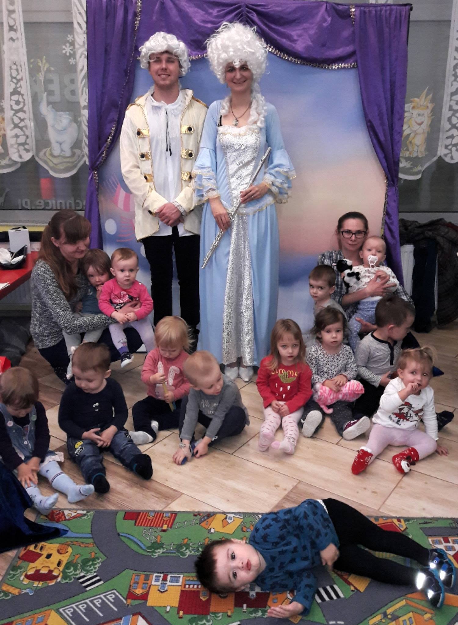 Dwoje aktorów w kostiumach z epoki, z perukami, pozuje z grupą małych dzieci siedzących na podłodze, na tle fioletowych zasłon i dekoracji scenicznej.