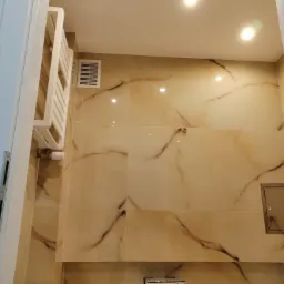 Wykończona łazienka z beżowymi płytkami imitującymi marmur, grzejnik drabinkowy i kratka wentylacyjna na ścianie, widok z dołu.