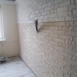 Dawid K - Ściana wykończona dekoracyjną cegłą w odcieniach bieli i beżu, z zamontowanym uchwytem do telewizora, widoczny grzejnik pod oknem i fragment podłogi.