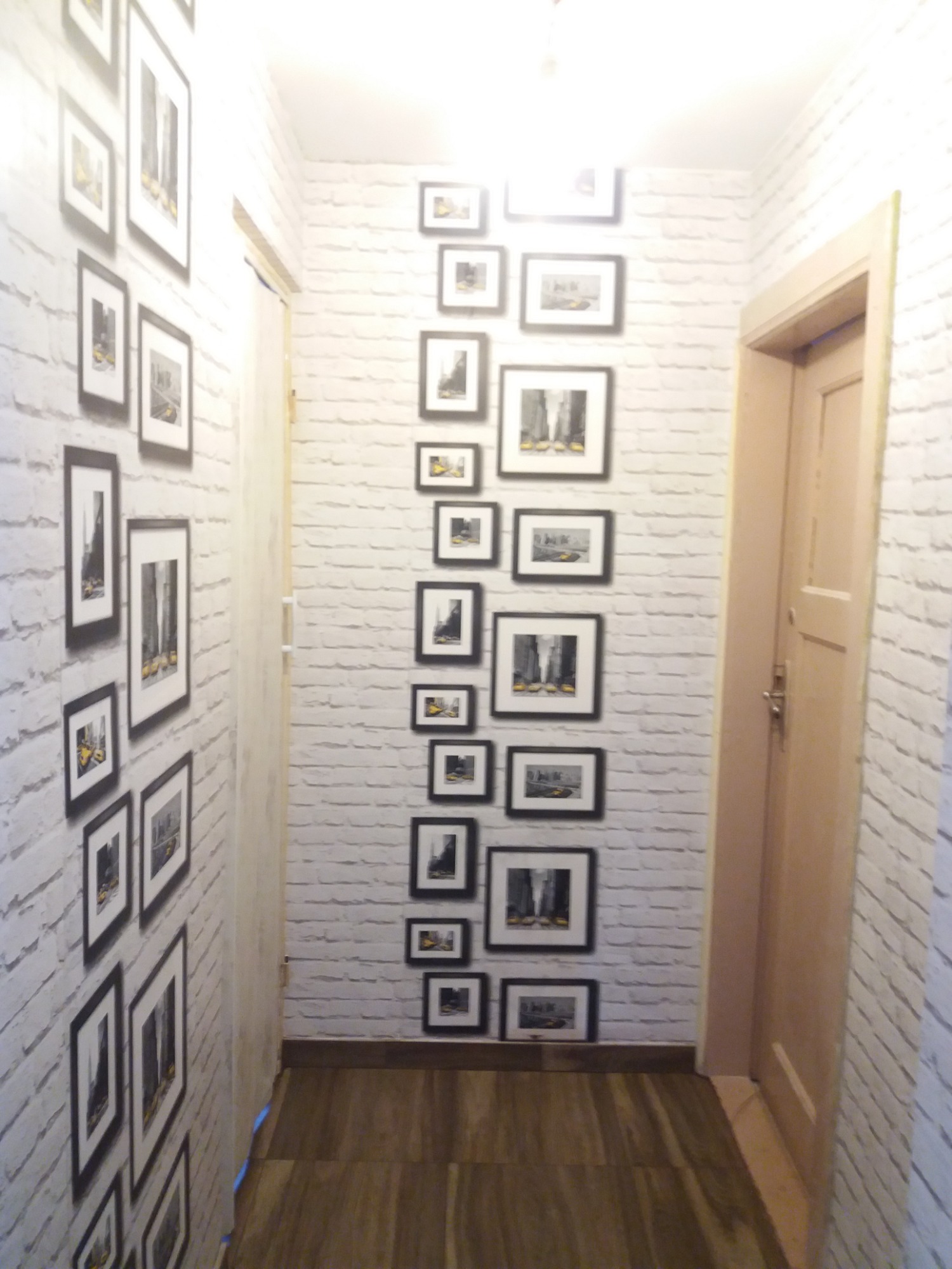 Wąski korytarz z drewnopodobną podłogą, ściany wykończone tapetą imitującą białą cegłę, z kolekcją czarno-białych zdjęć w czarnych ramkach na ścianach.