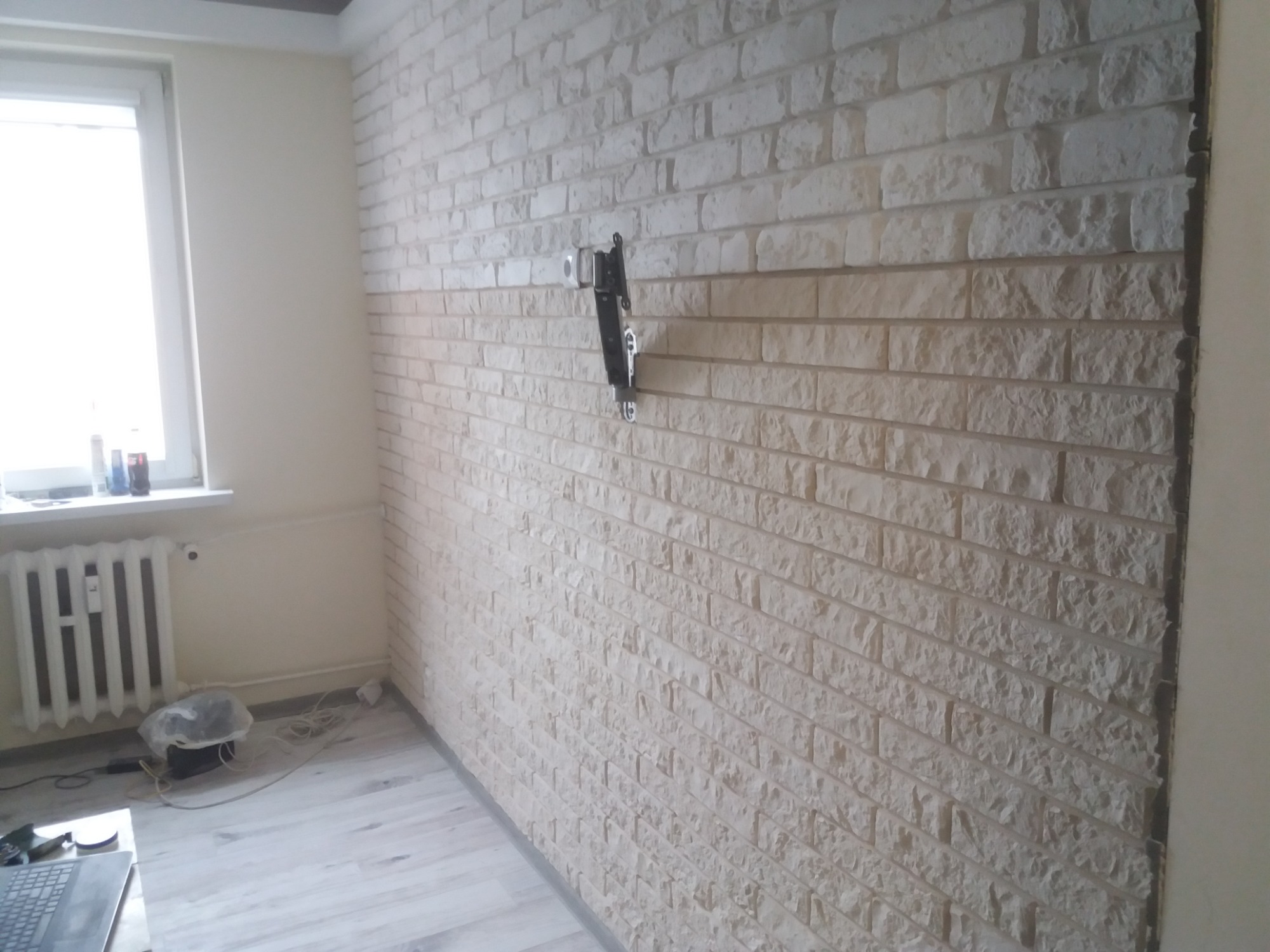 Ściana wykończona dekoracyjną cegłą w odcieniach bieli i beżu, z zamontowanym uchwytem do telewizora, widoczny grzejnik pod oknem i fragment podłogi.