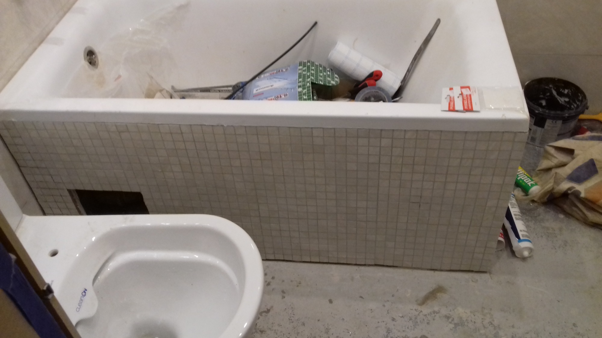 Niedokończona obudowa wanny mozaiką w łazience w trakcie remontu, widoczna toaleta, narzędzia i materiały budowlane w wannie.