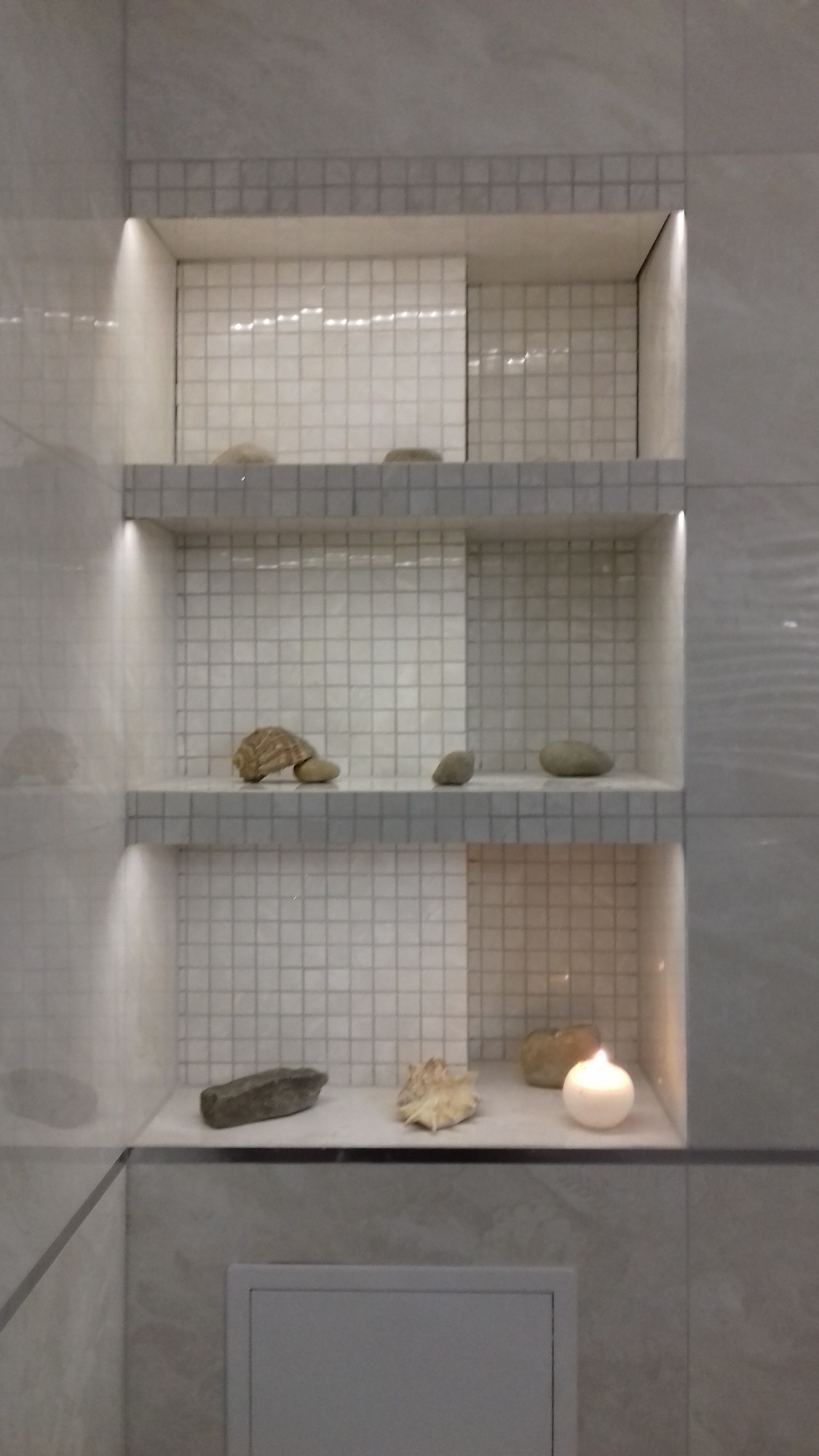 Wnęki w łazience wyłożone białą mozaiką, oświetlone od góry, z dekoracjami: kamieniami, muszlą i świecą.