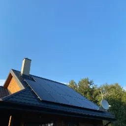 Panele fotowoltaiczne zamontowane na dachu drewnianego domu z kominem i anteną satelitarną, widok w słoneczny dzień.