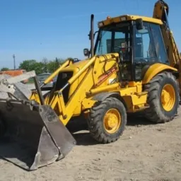 Żółta koparko-ładowarka stoi na piaszczystym placu, widoczny przedni łyżka z zębami i tylne ramię koparki, w tle drzewa i niebo.