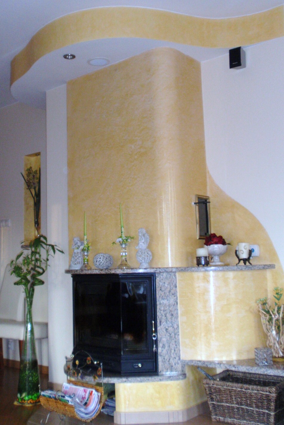 Wykończony salon z kominkiem obudowanym granitem, ściany pomalowane farbą strukturalną w odcieniach żółci, dekoracje na półce kominkowej.