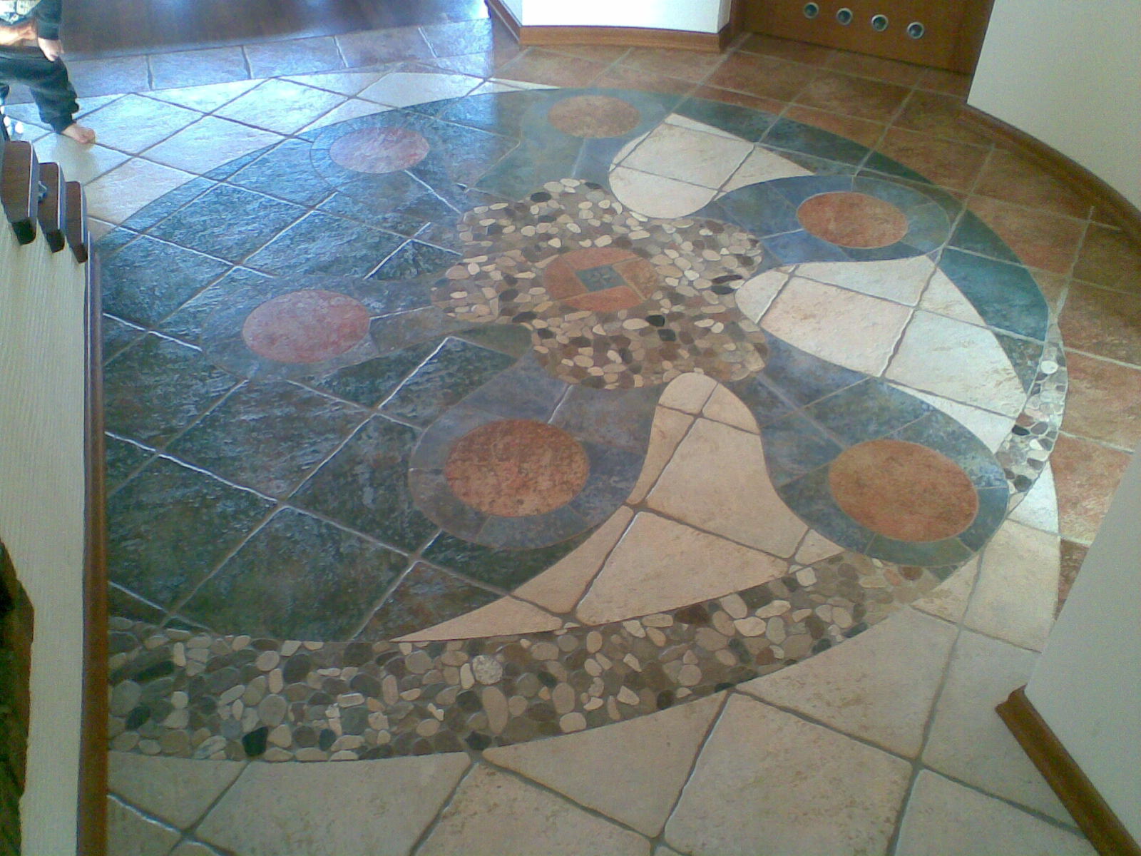 Oryginalna mozaika podłogowa z płytek i kamieni w kształcie yin-yang, z centralnym motywem kwiatowym z drobnych kamyków, otoczona geometrycznym wzorem z okrągłymi akcentami w odcieniach brązu i beżu.