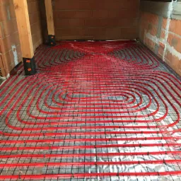 Czerwone rury ogrzewania podłogowego ułożone na folii izolacyjnej i siatce metalowej w surowym stanie, widoczne ceglane ściany i drewniane elementy konstrukcyjne poddasza.