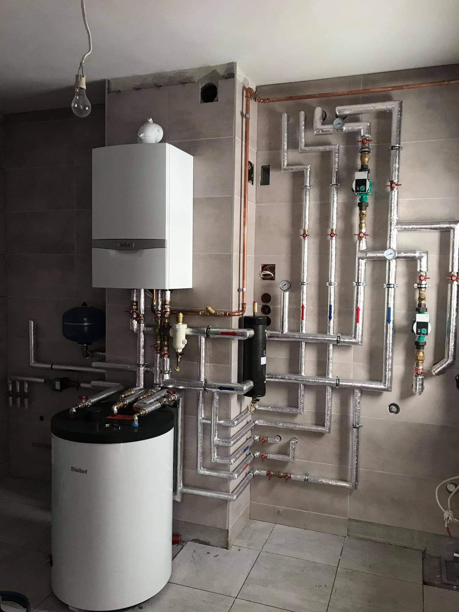Kompleksowa instalacja grzewcza w kotłowni z piecem gazowym Vaillant, zasobnikiem ciepłej wody, osprzętem i rurami w izolacji, z surową żarówką wiszącą na kablu.
