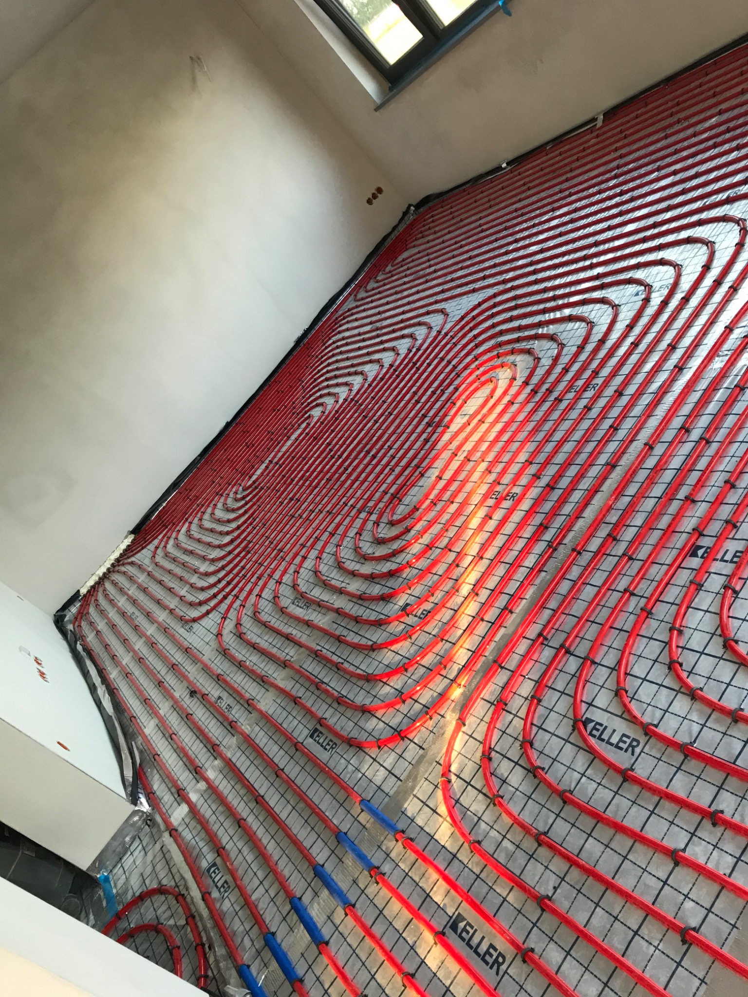 Czerwone rurki ogrzewania podłogowego ułożone na srebrnej folii z nadrukiem w kratkę, widok z góry na surową podłogę przed wylewką.