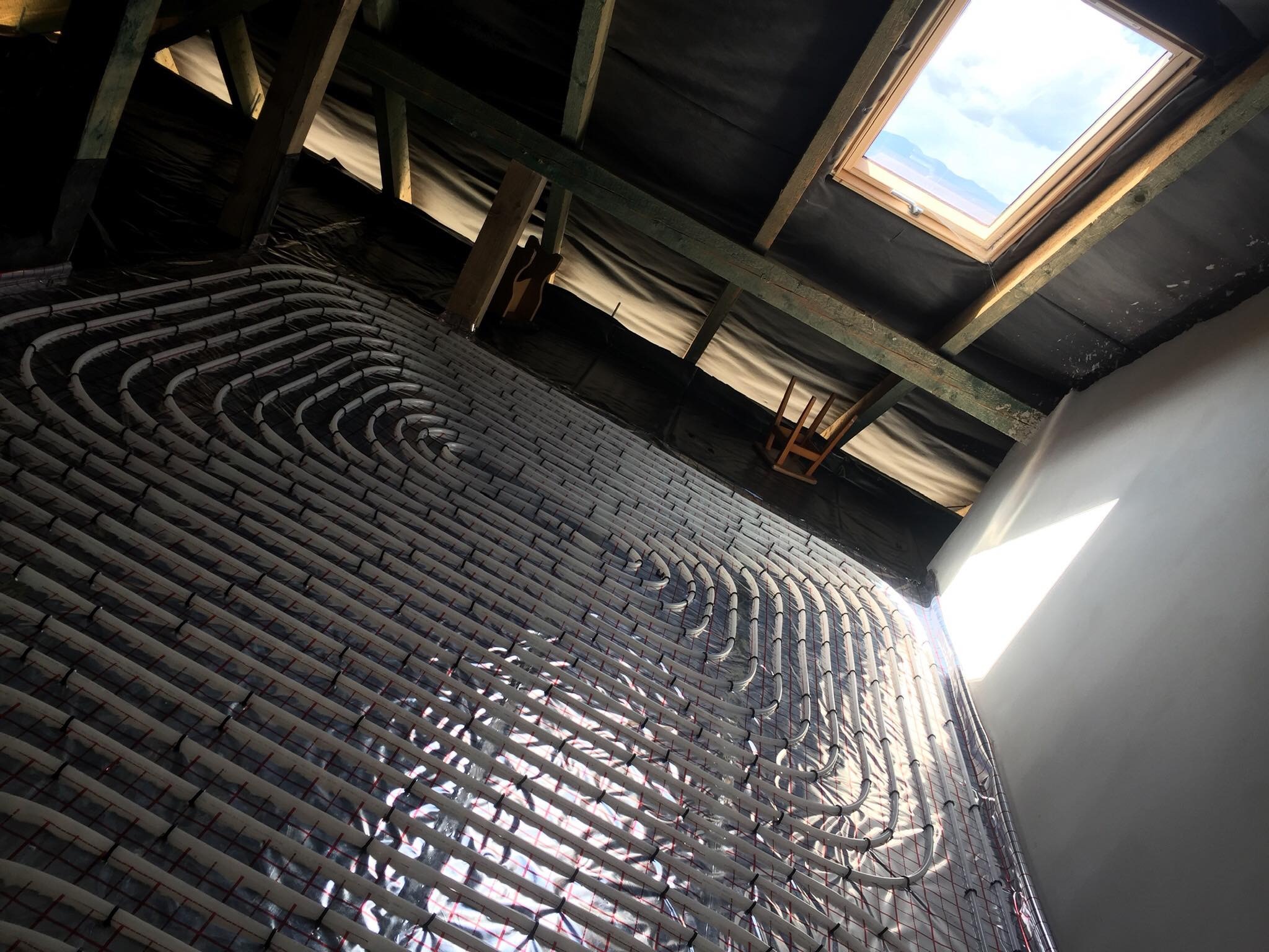 Montaż rur ogrzewania podłogowego na folii izolacyjnej w poddaszu, widoczne drewniane elementy konstrukcji dachu i okno dachowe.