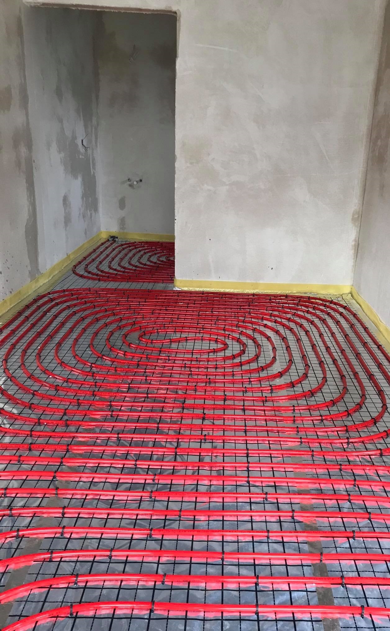Czerwone rury ogrzewania podłogowego ułożone na czarnej siatce w surowym pomieszczeniu z szarymi ścianami i żółtą taśmą brzegową.
