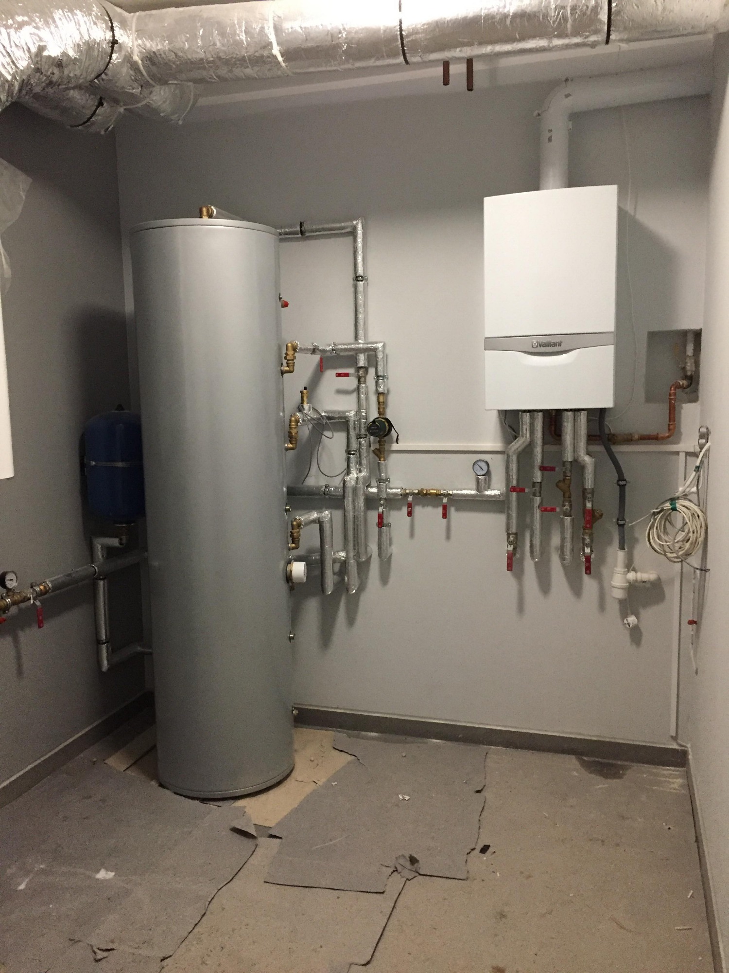 Nowoczesny, biały piec gazowy Vaillant, połączony rurami z szarym, pionowym zasobnikiem wody, zamontowany na szarej ścianie w pomieszczeniu z prowizorycznym podłożem.