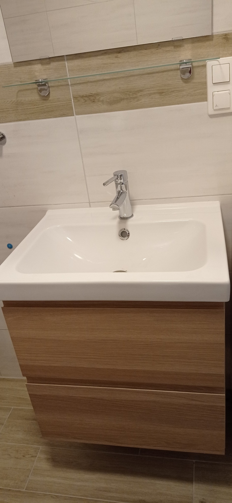 Biała umywalka z chromowaną baterią i szafką z drewnopodobnym frontem w łazience z jasnymi płytkami. Nad umywalką lustro z półką.