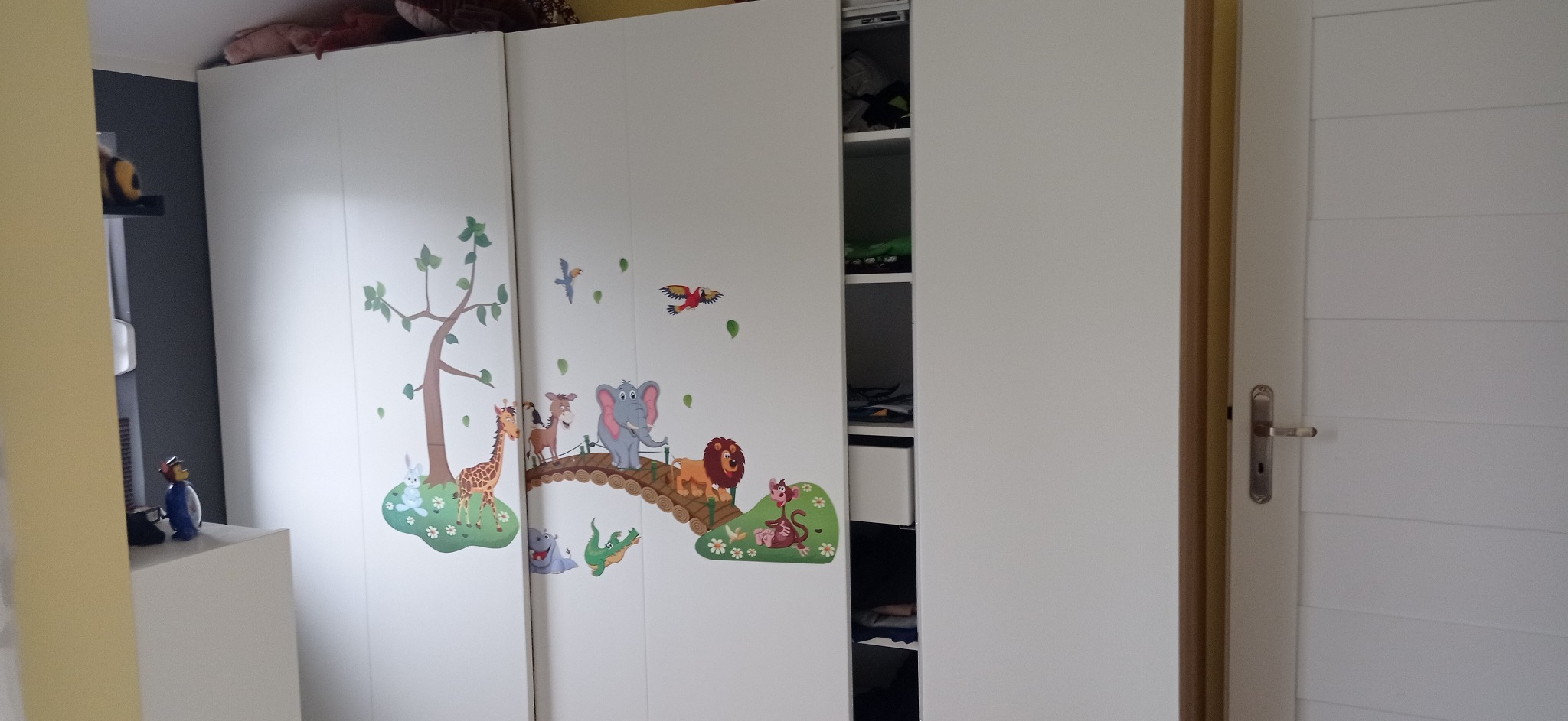 Biała szafa przesuwna z naklejkami zwierząt w pokoju dziecięcym. Częściowo otwarte drzwi ukazują półki z ubraniami. Widoczna także biała ściana i fragment drzwi.