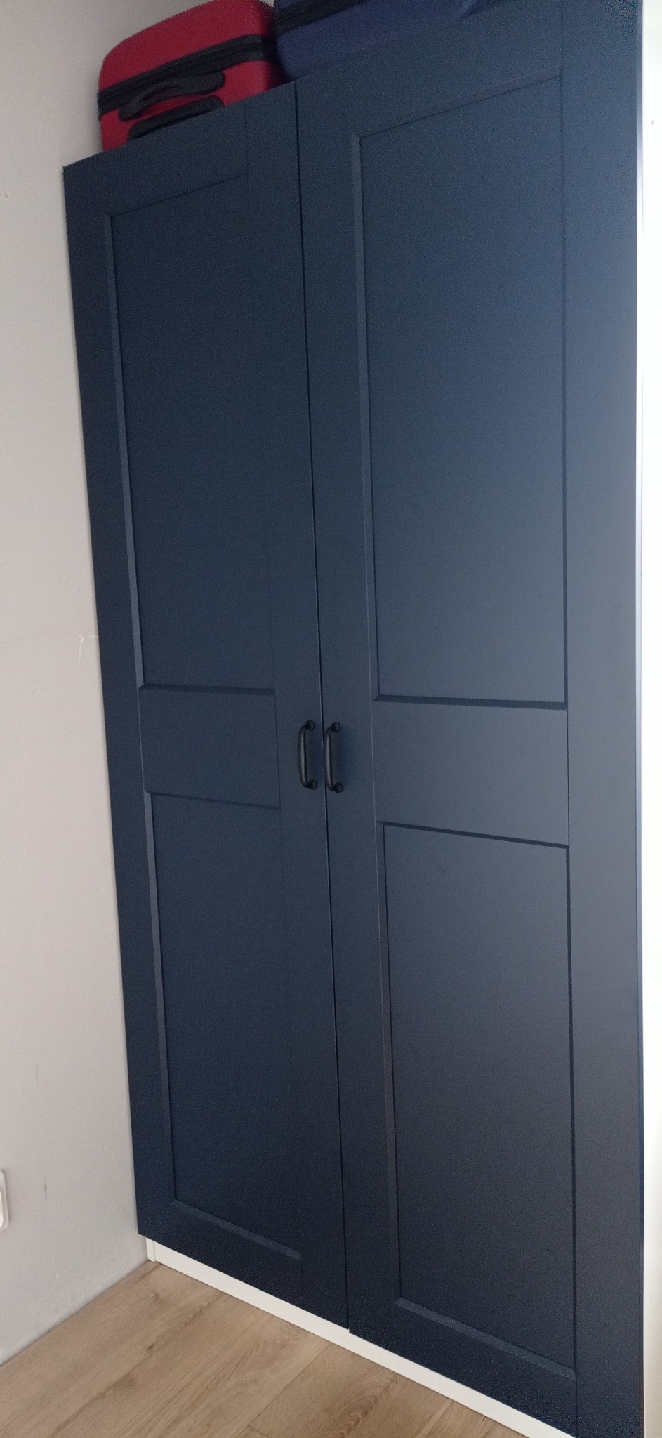 Granatowa, dwudrzwiowa szafa z czarnymi uchwytami, na której leżą dwie walizki, ustawiona na drewnianej podłodze przy białej ścianie.