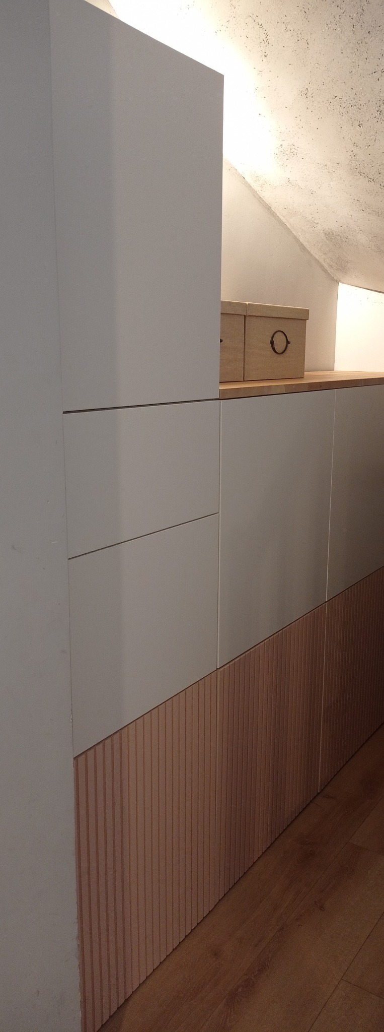 Biała zabudowa meblowa ze żłobionym panelem w dolnej części, dopasowana do skosu poddasza, z drewnianym blatem i pudełkami na nim.