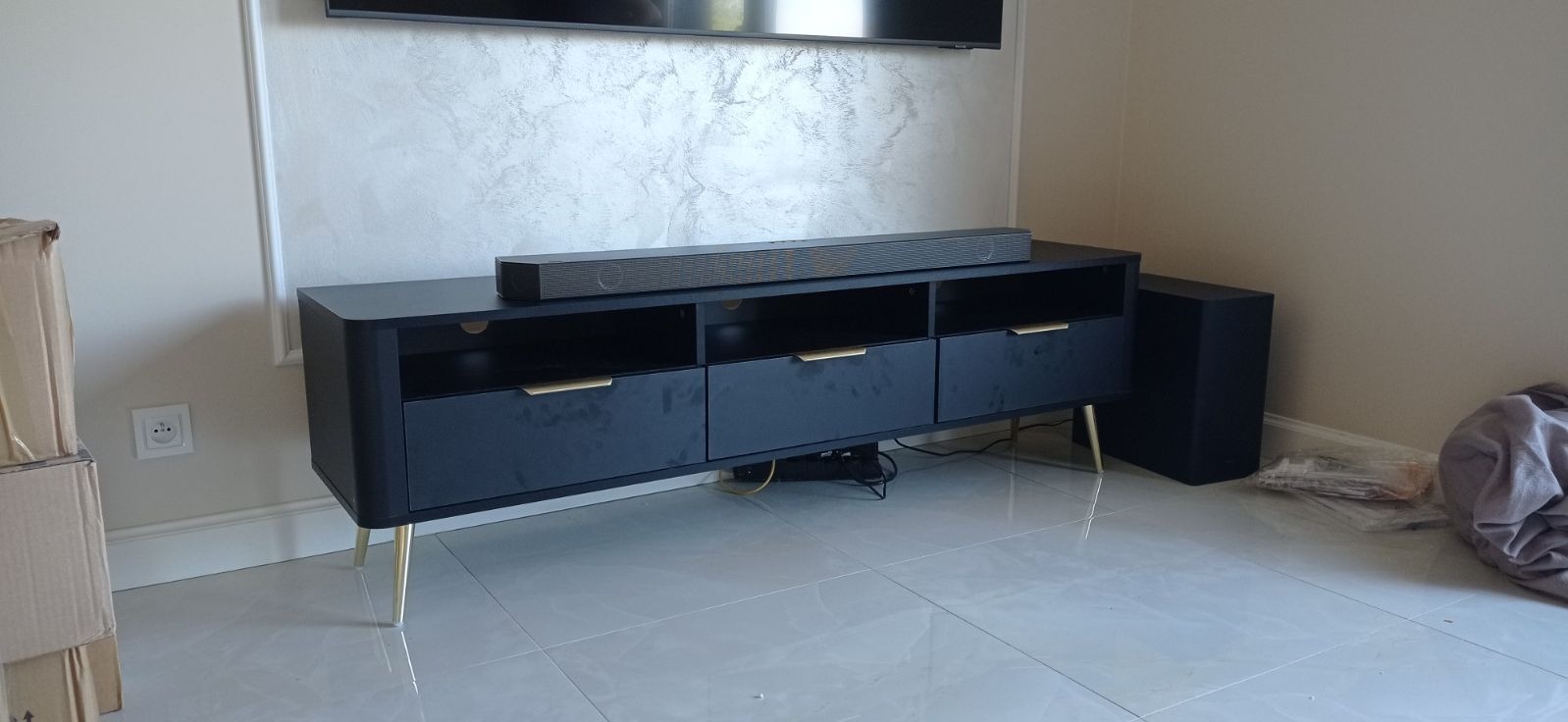 Nowoczesna, czarna szafka RTV z trzema szufladami i złotymi nóżkami, ustawiona na błyszczącej podłodze, z soundbarem na blacie i gładzią dekoracyjną w tle.