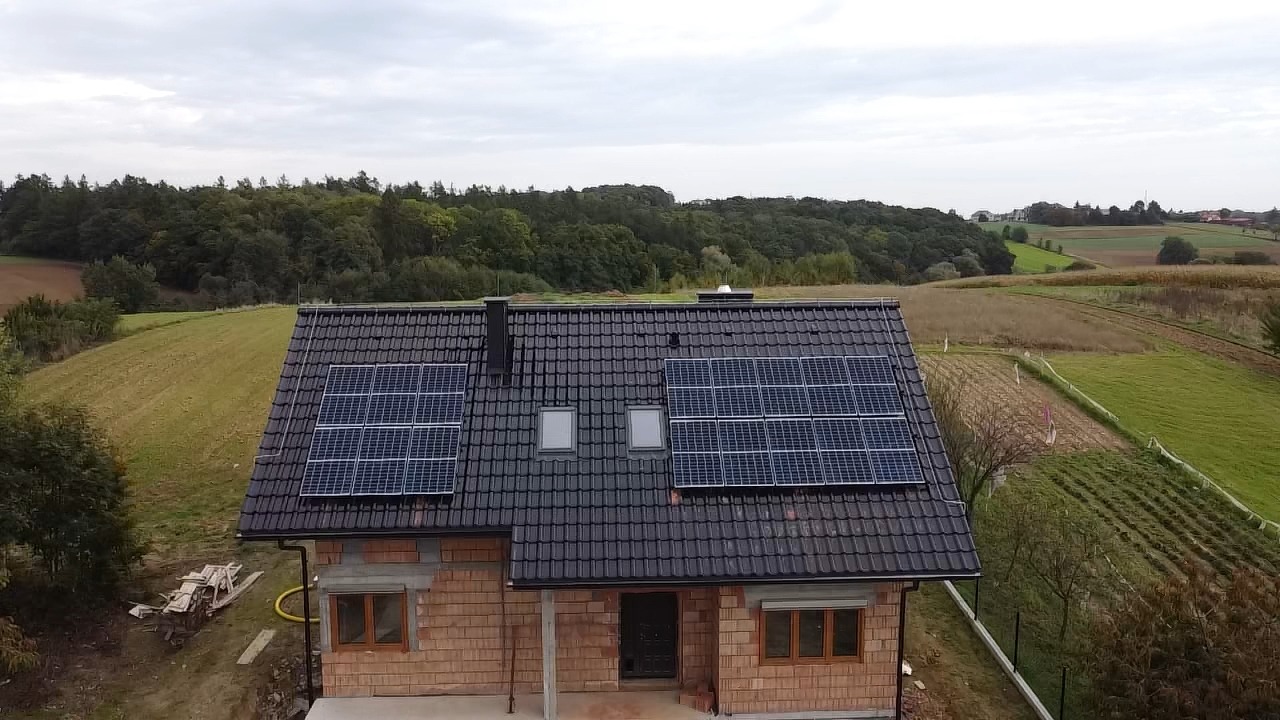 Widok z góry na dom jednorodzinny z ciemną dachówką i zamontowanymi panelami słonecznymi na dachu, w tle pola i las.