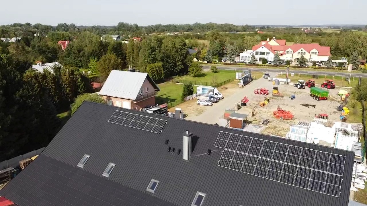 Widok z lotu ptaka na budynek z panelami słonecznymi na dachu, w tle zabudowania gospodarcze i sprzęt rolniczy.