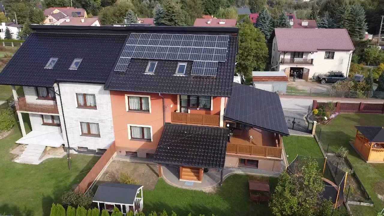 Widok z góry na dom z zainstalowanymi panelami słonecznymi na dachu, otoczony zielonym trawnikiem i drzewami.