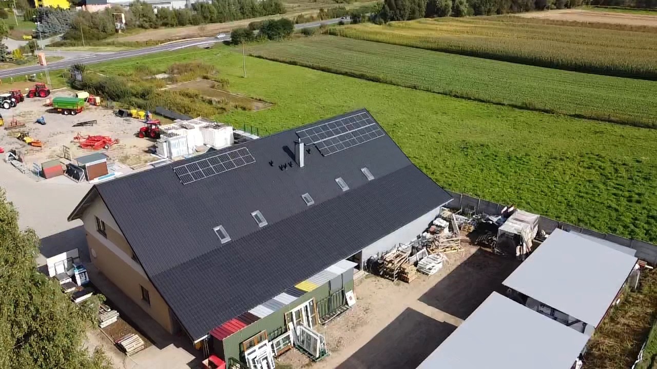 Widok z lotu ptaka na budynek z ciemnym dachem i panelami fotowoltaicznymi, otoczony zielonym polem i sprzętem rolniczym.