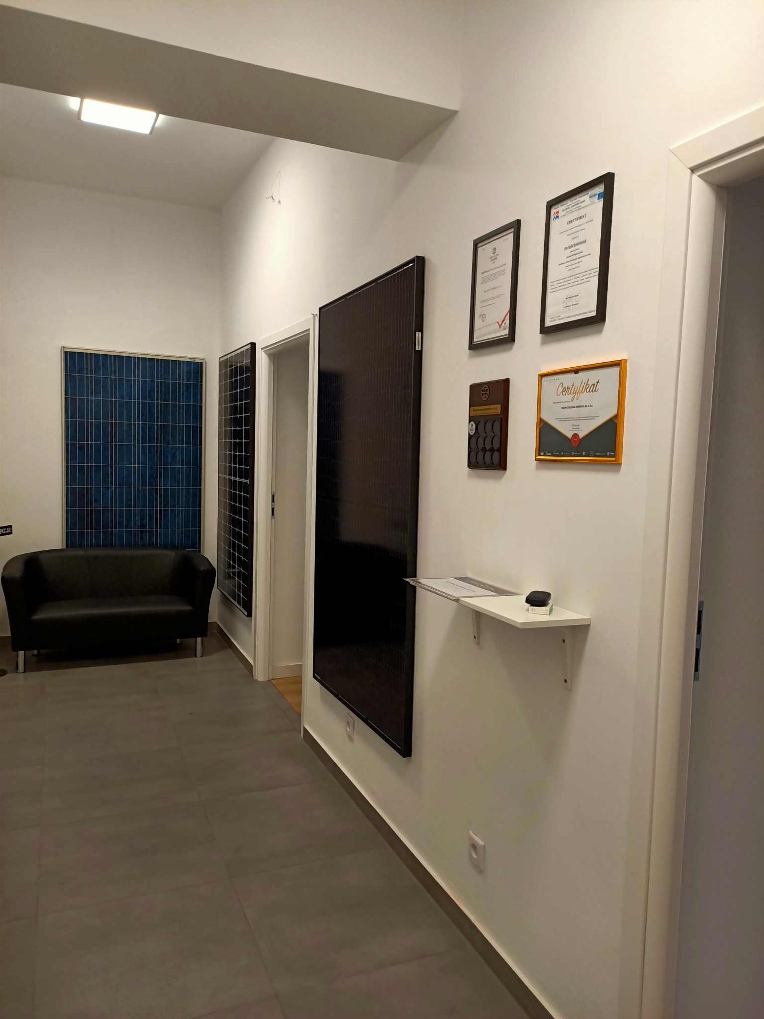 Wnętrze biura z ekspozycją paneli słonecznych, certyfikatami i dyplomami na ścianie, ukazujące różne typy i rozmiary paneli.