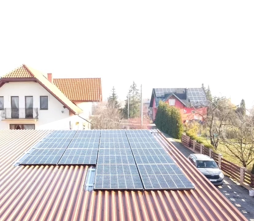 Panele słoneczne zamontowane na metalowym dachu domu jednorodzinnego, z widokiem na sąsiednie domy i zaparkowany samochód w słoneczny dzień.