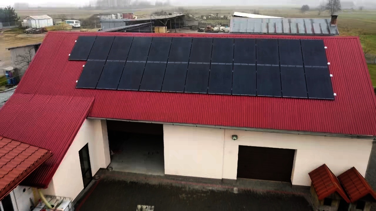 Instalacja paneli słonecznych na czerwonym dachu budynku gospodarczego, widok z góry w pochmurny dzień.