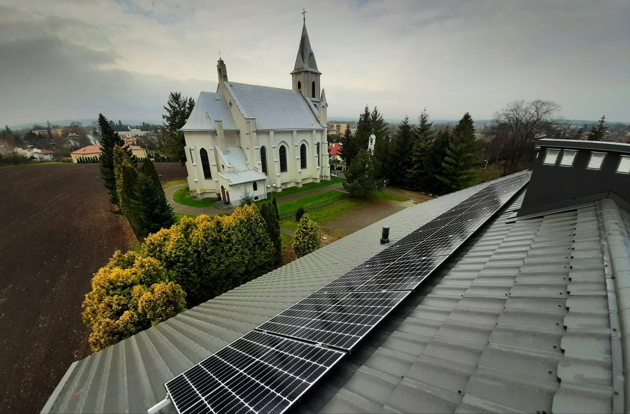 Panele słoneczne na szarym dachu, w tle kościół i zabudowa miejska w pochmurny dzień.