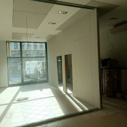 Ogolno budowlana - Wnętrze w trakcie montażu ścianki działowej z płyty gipsowo-kartonowej, widoczne profile metalowe i częściowo zamontowane płyty, zarys otworu drzwiowego, w tle okno z widokiem na ulicę i pracownika.